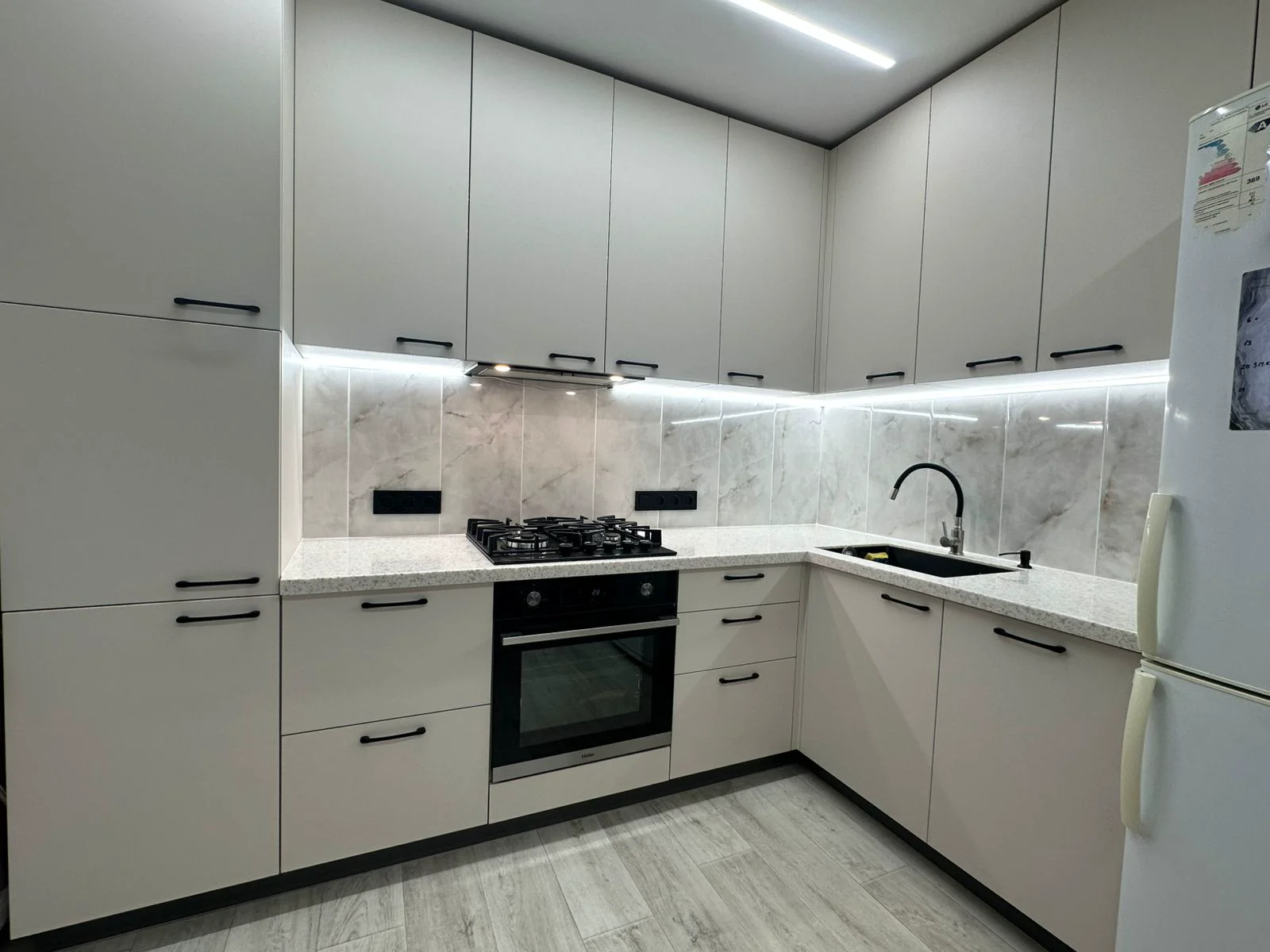 Белая угловая кухня с мраморным фартуком, чёрной фурнитурой и LED-подсветкой